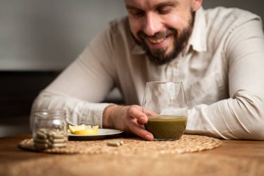 Kratom tüketim konsepti: erkek sakallı hippi elinde bir bardak yeşil sıvı, sıcak suda eritilmiş kratom tozu, kratom çayı, yakın çekim fotoğrafı, seçici odaklanma