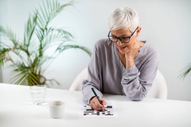 Engaging cognitive activity as a senior woman solves crossword puzzles, promoting mental stimulation and brain health clipart