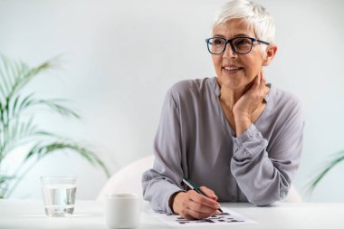 Engaging cognitive activity as a senior woman solves crossword puzzles, promoting mental stimulation and brain health clipart