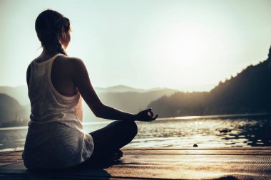 Lotus pozisyonunda bir kadın gün batımında göl kenarında yoga yapıyor.