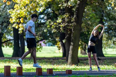 Yaz parkında ahşap engeller üzerinde egzersiz yapan çiftin görünüşü denge, koordinasyon ve fitness eğitimini birleştiriyor. Açık hava ortamında takım çalışması, sağlık ve aktif bir yaşam tarzı vurgulanıyor..