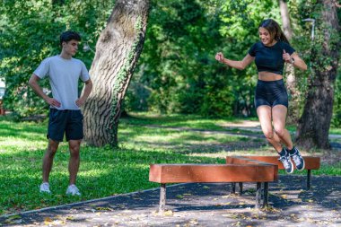 Parkta tahta kütüklerin üzerinden atlayan çift çeviklik, koordinasyon ve açık hava fitness aktivitesinde takım çalışması sergiliyor. Manzaralı park ortamı aktif ve sağlıklı bir yaşam tarzını vurguluyor.