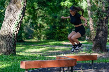 Parkta tahta kütüklerin üzerinden atlayan bir kadın açık hava fitness aktivitesinde çeviklik sergiliyor. Manzaralı park ortamı aktif ve sağlıklı bir yaşam tarzını vurguluyor.