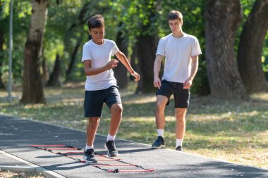 Genç bir çocuk, çeviklik merdiveni üzerinde bir antrenörle çeviklik tatbikatı yapıyor. Hız, koordinasyon ve uygunluğu geliştirmeye odaklanıyor. Çocuklar için spor eğitimi ve açık hava egzersizleri için mükemmel..