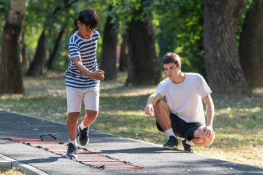 Oğlan dışarıda bir antrenörle çeviklik merdiveni talimi yapıyor, hız, koordinasyon ve uygunluğu geliştirmeye odaklanıyor. Açık hava düzeyi aktif olmayı vurguluyor 