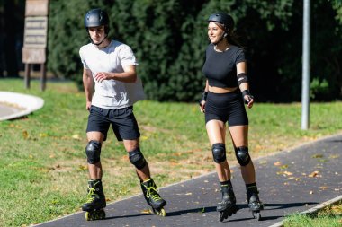 Genç çift manzaralı bir parkta birlikte paten kaymaktan hoşlanıyor. Çift yol boyunca süzülüyor, yeşilliklerle çevrili, eğlenceli ve aktif bir açık hava deneyimini paylaşıyor.