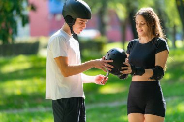 Adam kadının kaskına yardım ederken çift paten kaymaya hazırlanıyor. Bir anlık bakım ve takım çalışmasını paylaşıyorlar. Açık hava aktivitelerinin keyfini çıkarmadan önce güvenliği sağlıyorlar..