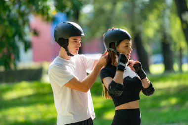 Adam kadının kaskına yardım ederken çift paten kaymaya hazırlanıyor. Bir anlık bakım ve takım çalışmasını paylaşıyorlar. Açık hava aktivitelerinin keyfini çıkarmadan önce güvenliği sağlıyorlar..