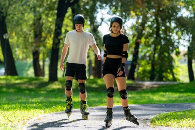 Genç çift, güneşli bir yaz gününde yemyeşil bir parkta paten kaymaktan hoşlanıyor. Çift, etkin bir yaşam tarzını kucaklayarak ve keyifli bir açık hava deneyimini paylaşarak manzaralı yollarda süzülür..