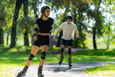 Genç çift, güneşli bir yaz gününde yemyeşil bir parkta paten kaymaktan hoşlanıyor. Çift, etkin bir yaşam tarzını kucaklayarak ve keyifli bir açık hava deneyimini paylaşarak manzaralı yollarda süzülür..