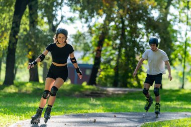 Genç çift, güneşli bir yaz gününde yemyeşil bir parkta paten kaymaktan hoşlanıyor. Çift, etkin bir yaşam tarzını kucaklayarak ve keyifli bir açık hava deneyimini paylaşarak manzaralı yollarda süzülür..