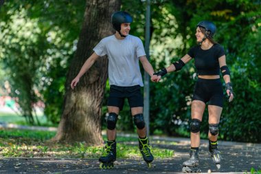 Genç çift, güneşli bir yaz gününde yemyeşil bir parkta paten kaymaktan hoşlanıyor. Çift, etkin bir yaşam tarzını kucaklayarak ve keyifli bir açık hava deneyimini paylaşarak manzaralı yollarda süzülür..