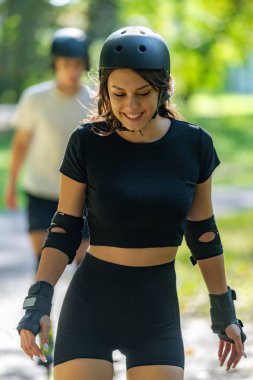 Genç çift, güneşli bir yaz gününde yemyeşil bir parkta paten kaymaktan hoşlanıyor. Çift, etkin bir yaşam tarzını kucaklayarak ve keyifli bir açık hava deneyimini paylaşarak manzaralı yollarda süzülür..