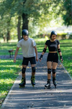 Genç çift parkta birlikte paten kayıyor, aktif bir açık hava aktivitesinin tadını çıkarıyor, fitness, eğlence ve bereketli yeşillik ve temiz hava ortamında kaynaşmayı teşvik ediyor..