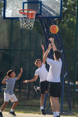 Çocuklar açık hava basketbol antrenmanında koçlarıyla takılıyor, sahada top sürme, pas atma ve atış becerilerini parlak doğal ışık altında geliştirerek, takım çalışmasını ve koordinasyonunu teşvik ederek geliştiriyorlar..