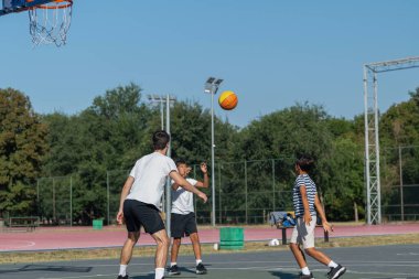 Çocuklar açık hava basketbol antrenmanında koçlarıyla takılıyor, sahada top sürme, pas atma ve atış becerilerini parlak doğal ışık altında geliştirerek, takım çalışmasını ve koordinasyonunu teşvik ederek geliştiriyorlar..