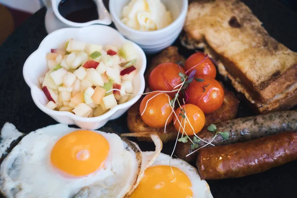 Full plate of traditional English breakfast - fried eggs, toasted bread, grilled cherry tomato, sausage, beans and fruit salad with marple syrup