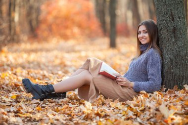 Sonbahar ormanında yaprakların üzerinde oturup kitap okuyan genç bir kadın.