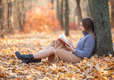 Sonbahar ormanında yaprakların üzerinde oturmuş kitap okuyan genç bir kadın.