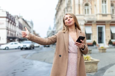 Sarışın kadın araba yakalıyor, Avrupa şehrinde el hareketleriyle akıllı telefon tutuyor. Taksi çağır.