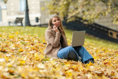 Genç bayan serbest çalışan, düşen yaprakların üzerinde oturuyor dizüstü bilgisayarla çalışıyor ve sonbahar parkında telefonla konuşuyor. Serbest çalışma, uzak çalışma veya eğitim