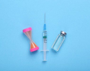 Mavi zemin üzerinde şırınga ve kum saati olan aşı şişesi.