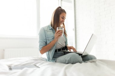 Genç hippi kadın yatakta dizüstü bilgisayar kullanıyor ve odada kahve içiyor. Uzak çalışma veya eğitim, çevrimiçi sohbet, serbest çalışma