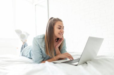 Genç gülümseyen kadın, aydınlık bir günde odada yatarken dizüstü bilgisayar kullanıyor.