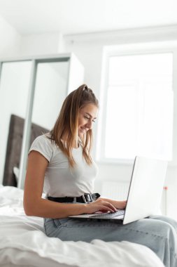 Genç gülümseyen kadın, aydınlık odada yatarken dizüstü bilgisayar kullanıyor.