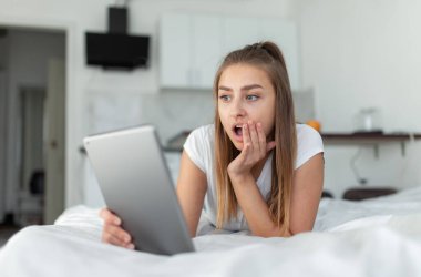 Yatakta yatarken tabletten video izleyen şaşırmış kadın.