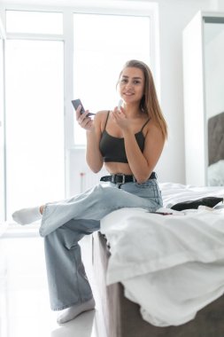 Ruj ve aynayla gülümseyen kadın aydınlık odada yatağında oturuyor.