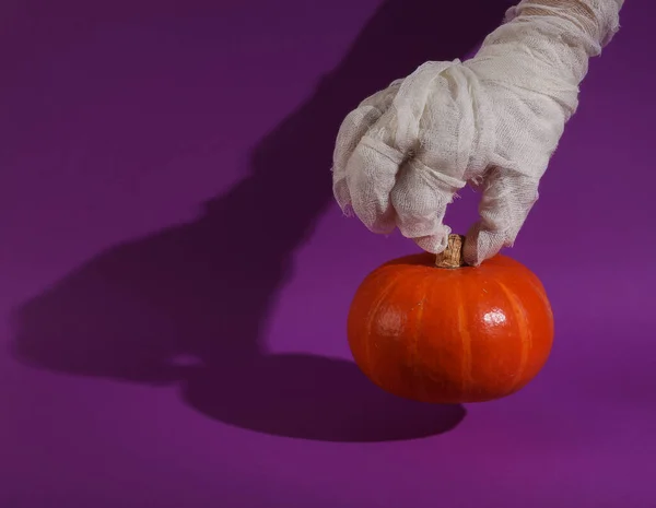 Mummy\'s hand wrapped in white bandages holds pumpkin on purple background with shadow. Happy Halloween. Trick or Treat