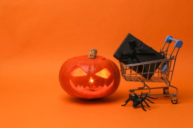 Cadılar Bayramı balkabağı kabağı kabağı feneri yanan mumlar ve turuncu arka planda siyah hediye kutusu olan alışveriş arabası.