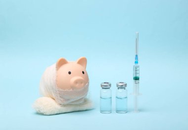 Tedavi konsepti. Bandaj ve şırıngaya sarılı domuz kumbarası ve mavi arka planda aşı şişeleri.