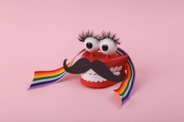 Funny toy clockwork jumping teeth with eyes and false eyelashes, mustache, rainbow tape on pink background. Transgender, indeterminate gender concept