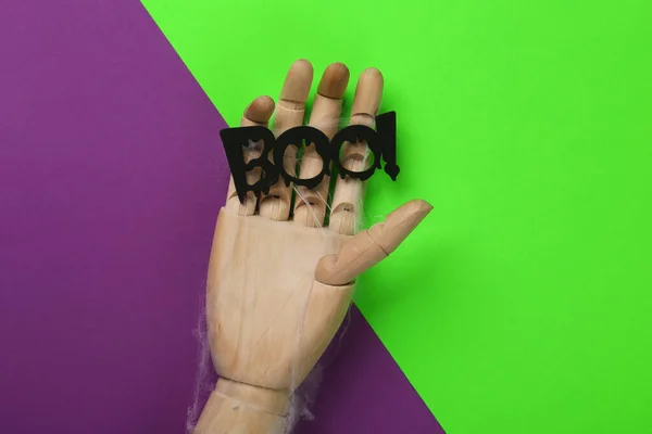 Halloween minimal still life. Wooden hand holding boo! word on purple green background. Top view