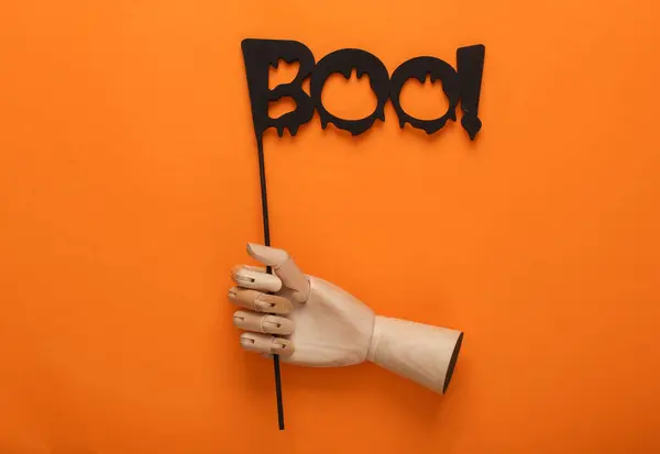 halloween layout. Wooden puppet hand holding the word boo! on a stick, orange background. Creative layout