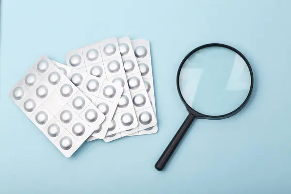 Metal blisters of pills with magnifying glass on blue background. Search for medicines concept