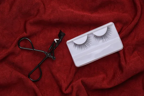 Professional eyelash curler with false eyelashes on red crumpled silk background. Top view