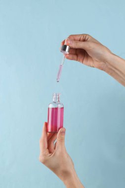 Bottle of face serum with dropper in female hands on blue background.