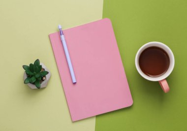 Pink notepad or diary with a cup of coffee and a plant on a green background. Flat lay