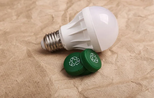 Plastic bottle caps with circular recycling symbol and light bulb on crumpled craft paper background. Round recycle concept