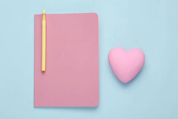 Girl's diary with a pink blank cover and a heart on a blue background. Top view, flat composition