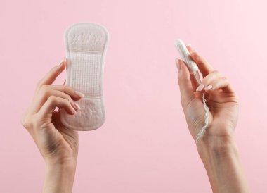 Kadının eli pembe arka planda tampon ve ped tutuyor. Kadın hijyeni, adet görme, kritik günler.