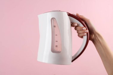 Kadının eli pembe arka planda elektrikli su ısıtıcısını tutuyor.