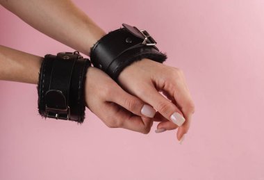 Pembe arka planda deri kelepçeli kadın elleri. BDSM aksesuarlar, hakimiyet, seks oyunları
