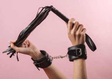 Deri kelepçeli kadın elleri pembe arka planda deri bir kamçı tutuyor. BDSM aksesuarlar, hakimiyet, seks oyunları