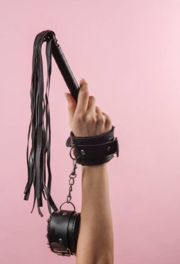 Deri kelepçeli kadın eli pembe arka planda deri kamçı tutuyor. BDSM aksesuarlar, hakimiyet, seks oyunları