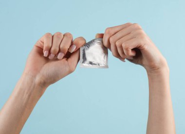 Kadın eli mavi arka planda bir prezervatif paketini açıyor.