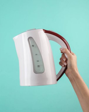 Kadının eli mavi arka planda elektrikli su ısıtıcısını tutuyor.